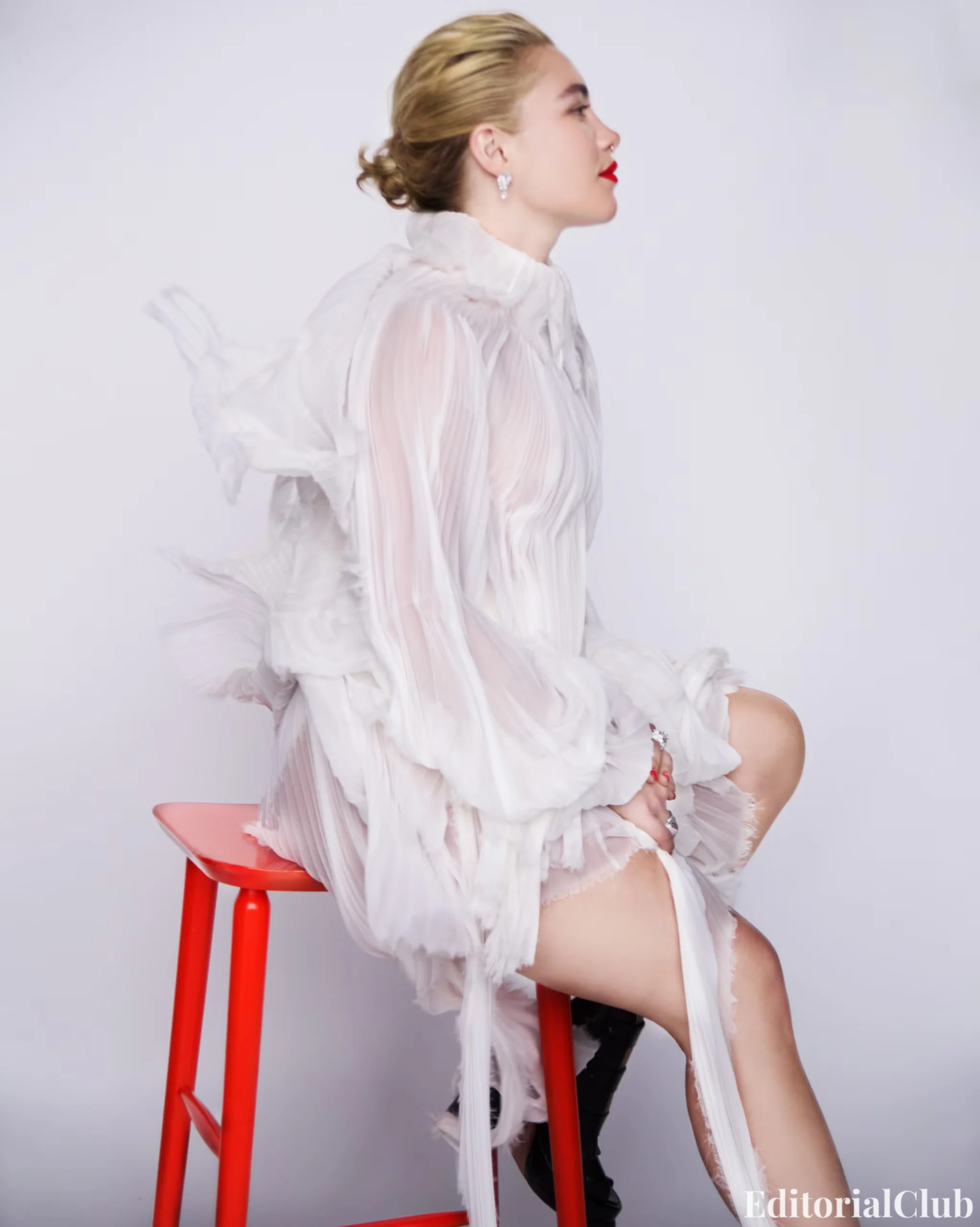 Florence Pugh sitting on a red stool in a sheer white pleated dress with slicked-back hair.