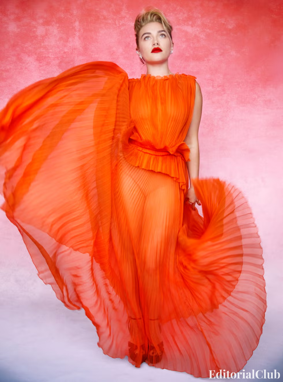 Florence Pugh in a flowing sheer orange gown, standing powerfully in front of a textured pink background.