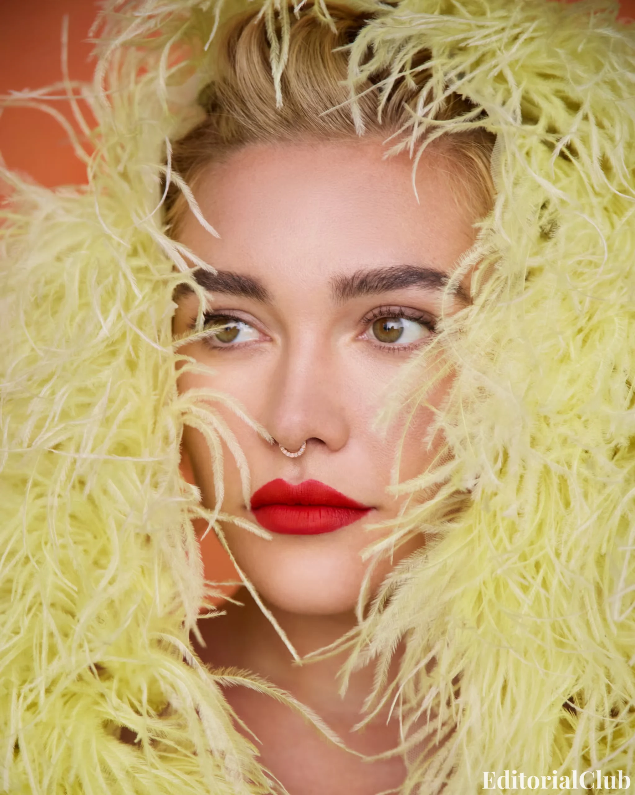 Close-up of Florence Pugh’s face framed by soft yellow feathers, highlighting red lips and a diamond nose ring.