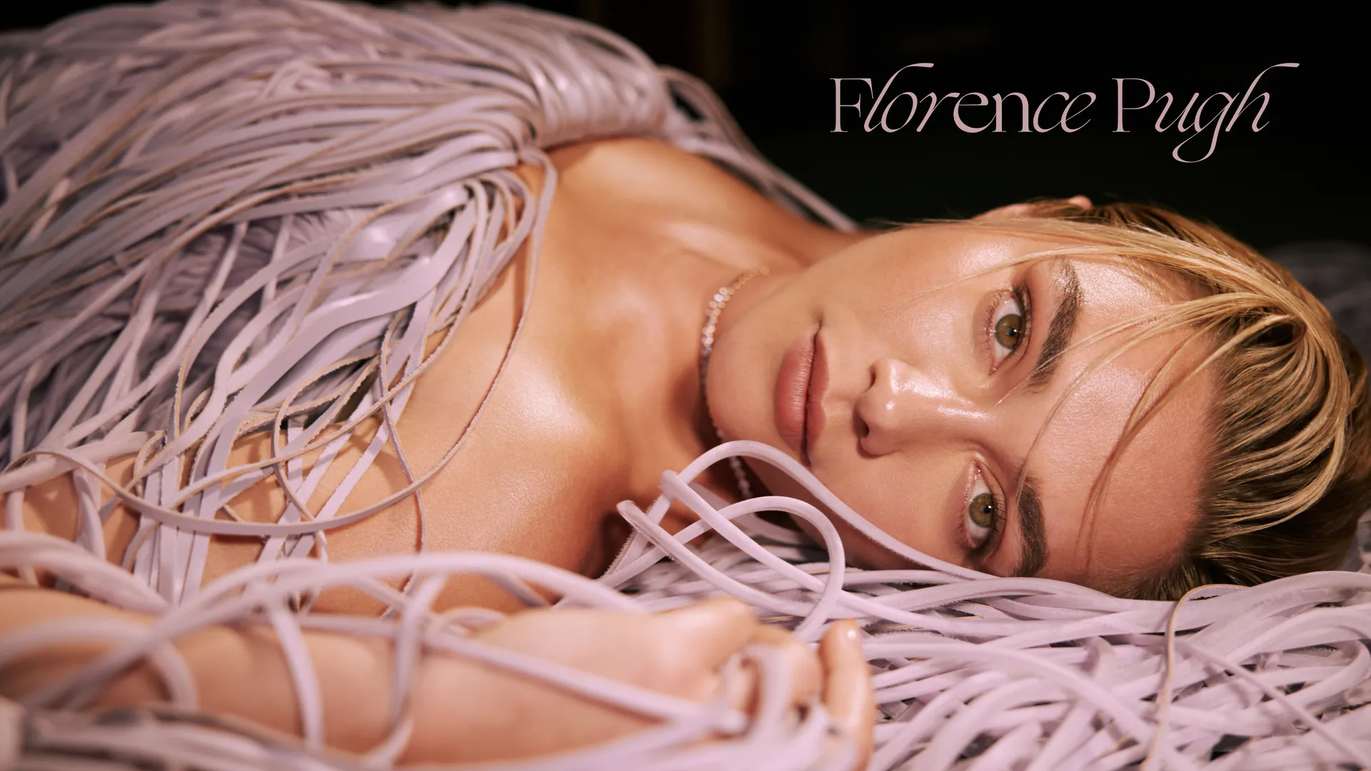 Florence Pugh lying down in a lavender fringe dress, gazing into the camera with dramatic lighting – Who What Wear May 2025.