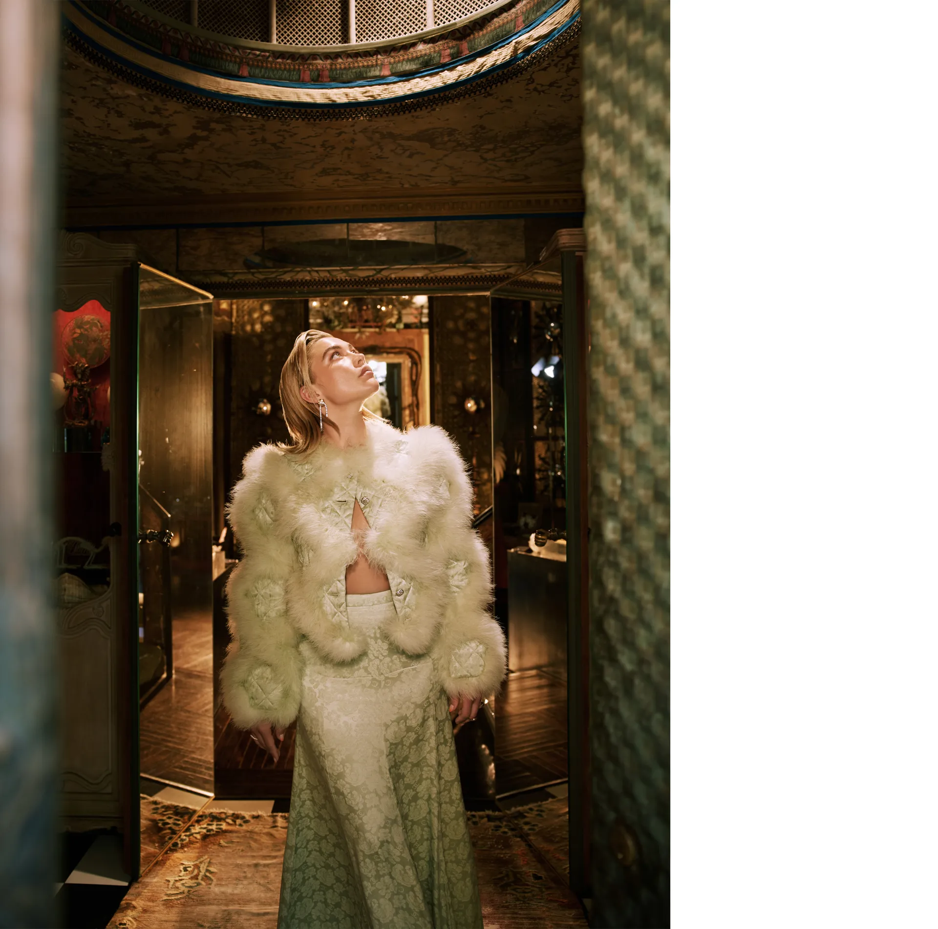 Florence Pugh gazing upward in an embellished white fur jacket and brocade skirt inside a richly decorated room – Who What Wear May 2025 photoshoot.