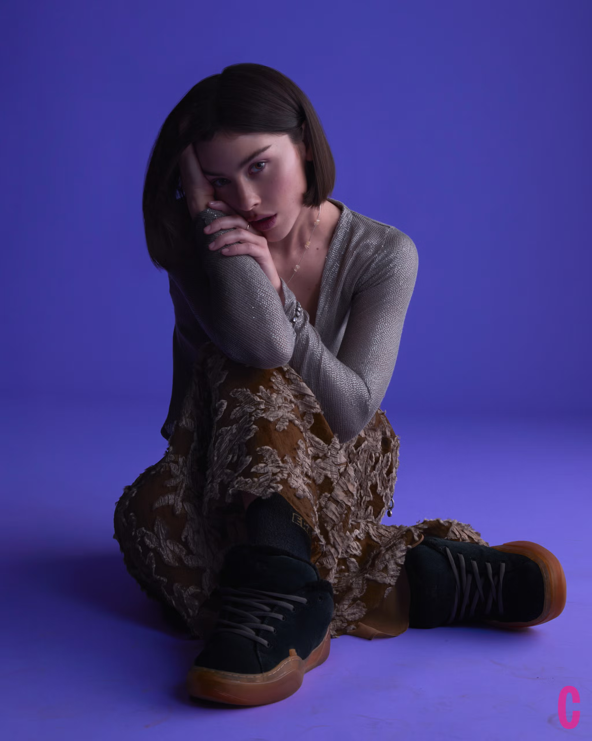 Gracie Abrams sits cross-legged in a metallic top, floral textured skirt, and black sneakers during her Cosmopolitan February 2025 shoot.