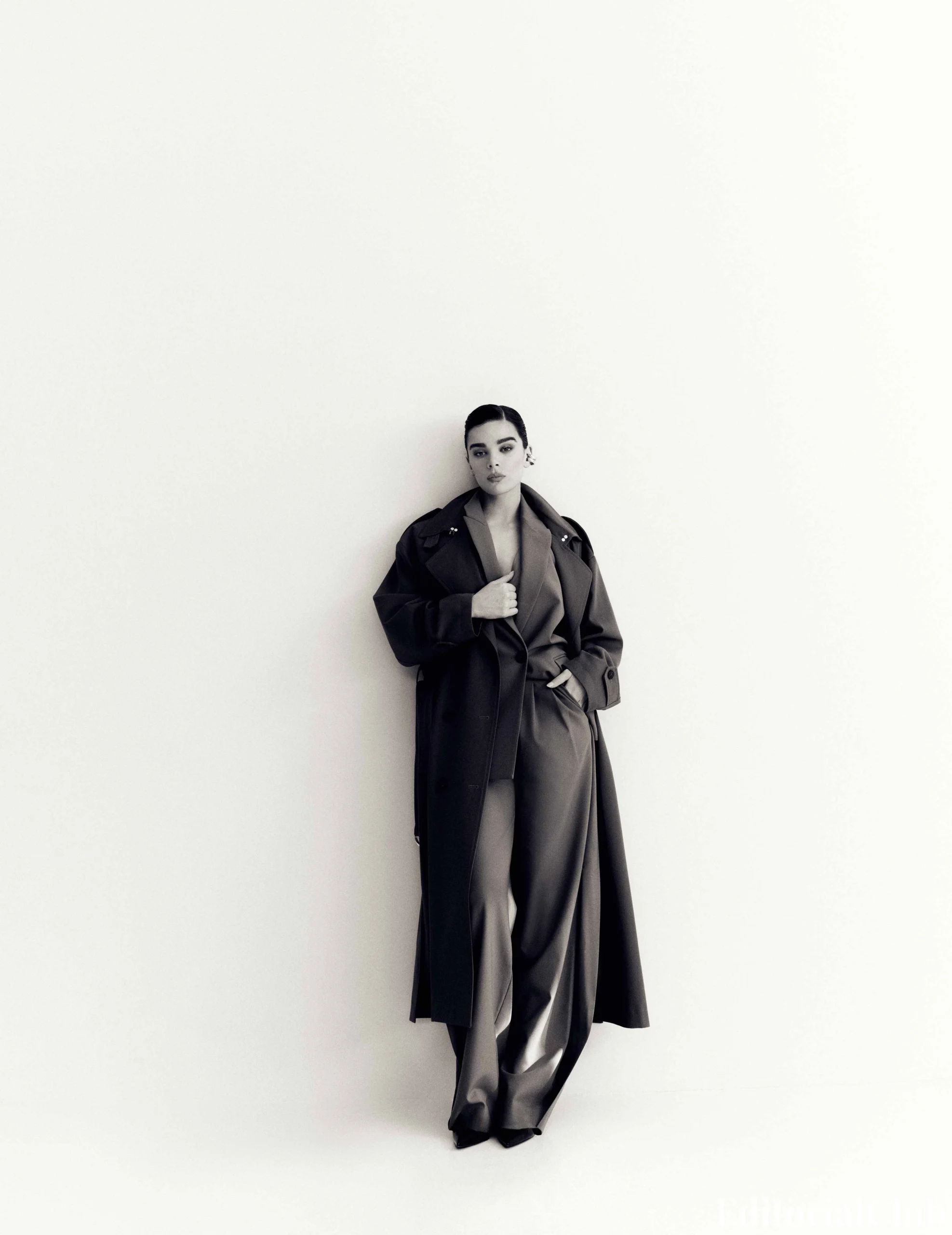Full-body shot of Hailee Steinfeld leaning against a wall in a long dark coat and pants for Flaunt 2025.