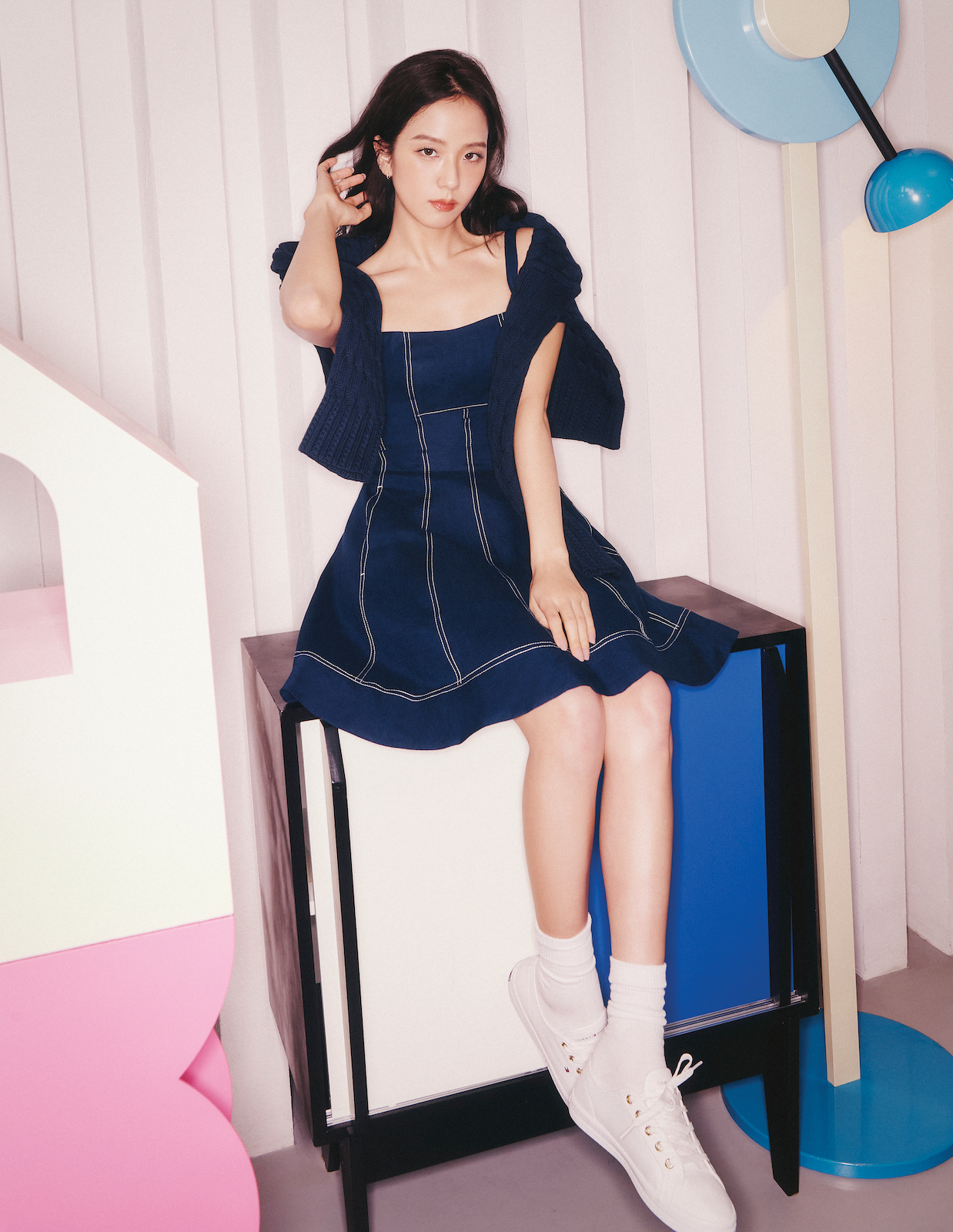 Jisoo sits elegantly in a dark denim dress with contrast stitching and a navy sweater draped over her shoulders, set against a geometric, pastel-toned room.