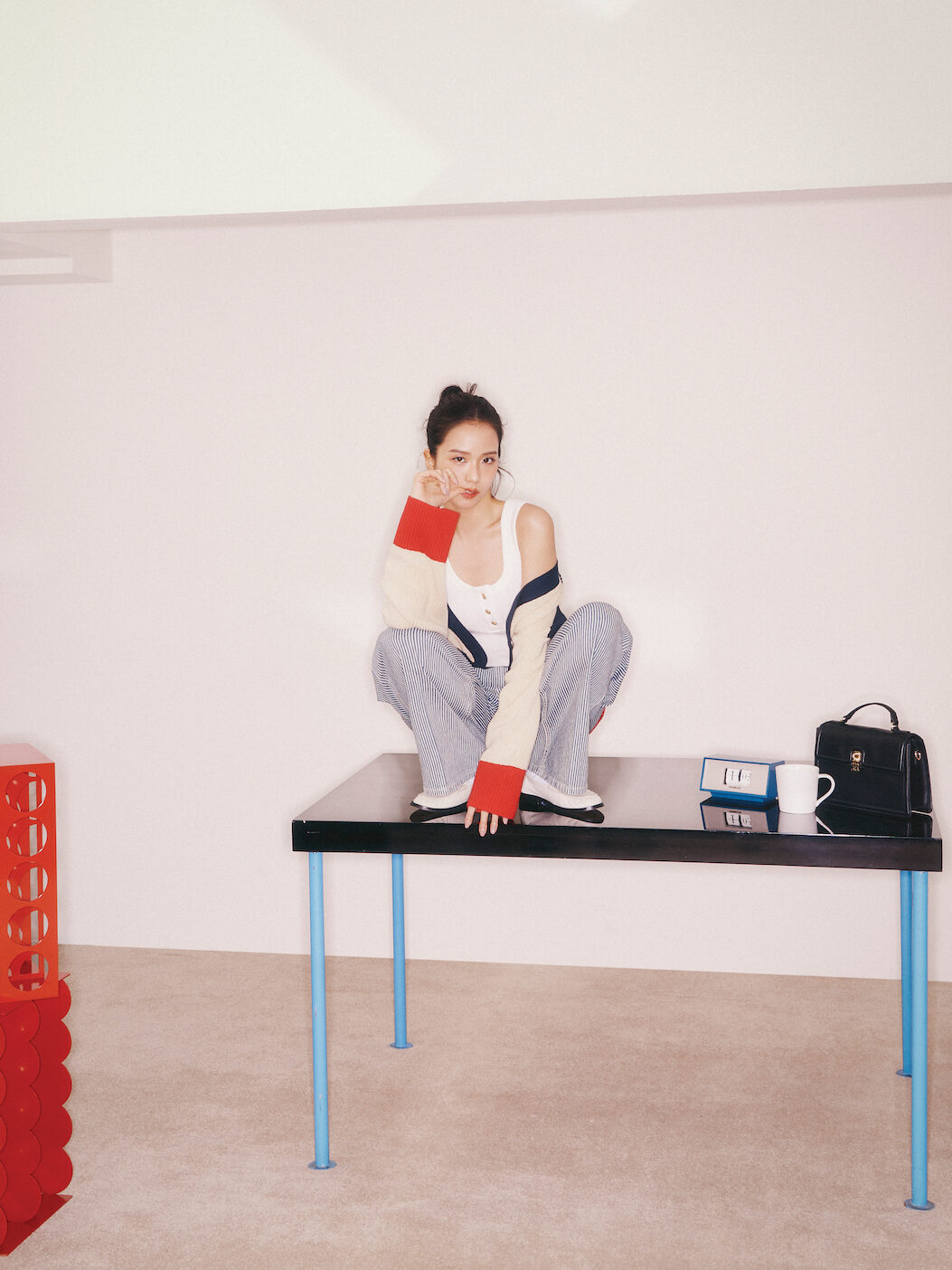 Jisoo crouches playfully on a minimalist black table, wearing pinstripe pants, a white tank, and a cozy striped cardigan with bold red cuffs.