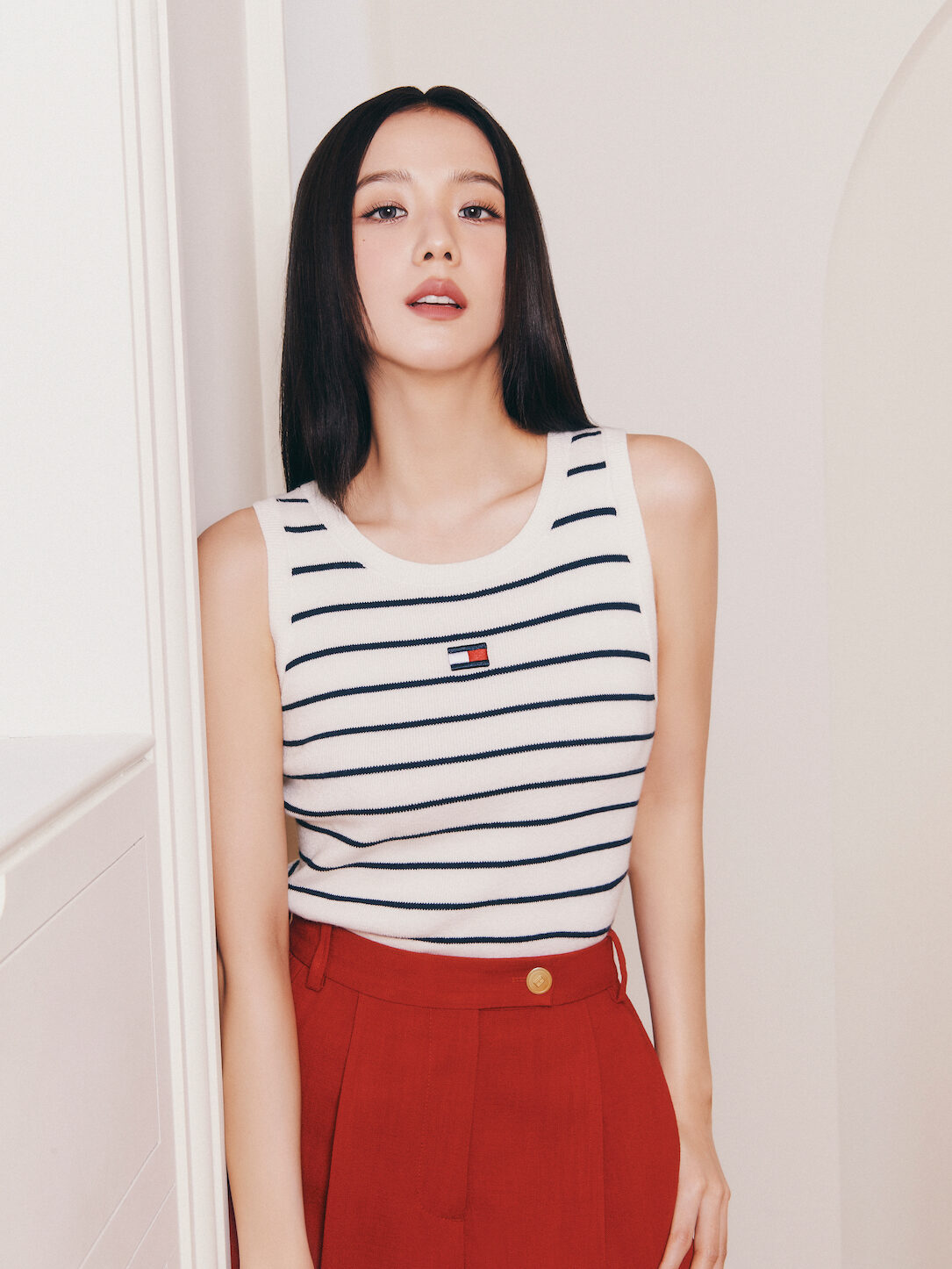 Jisoo poses in a sleeveless white tank with navy stripes and high-waisted red pleated trousers, styled with a clean and modern aesthetic in a white interior.