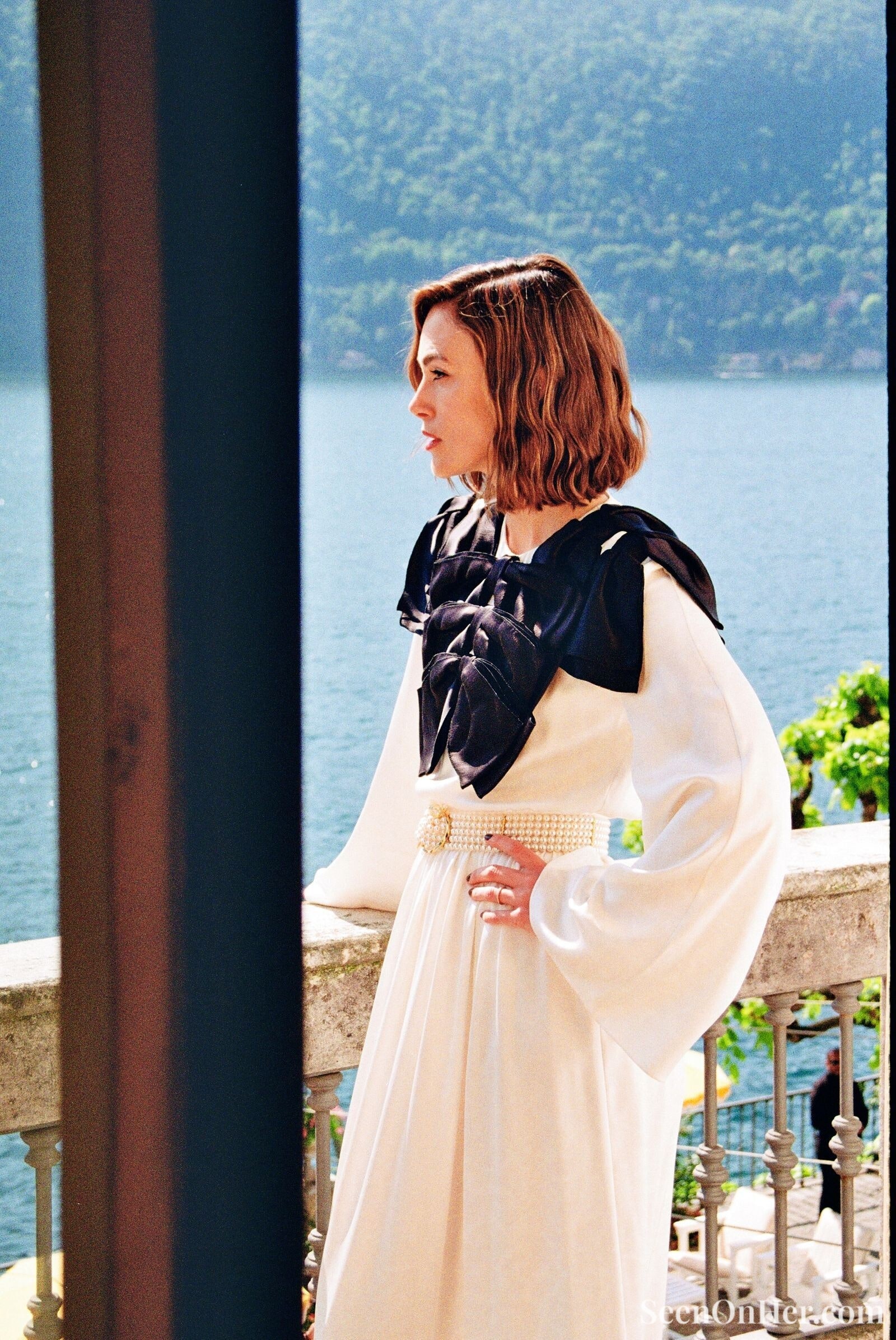 Side profile of Keira Knightley wearing a white gown with a pearl belt and black satin bow embellishments, standing on a lakeside balcony with scenic mountains behind her.
