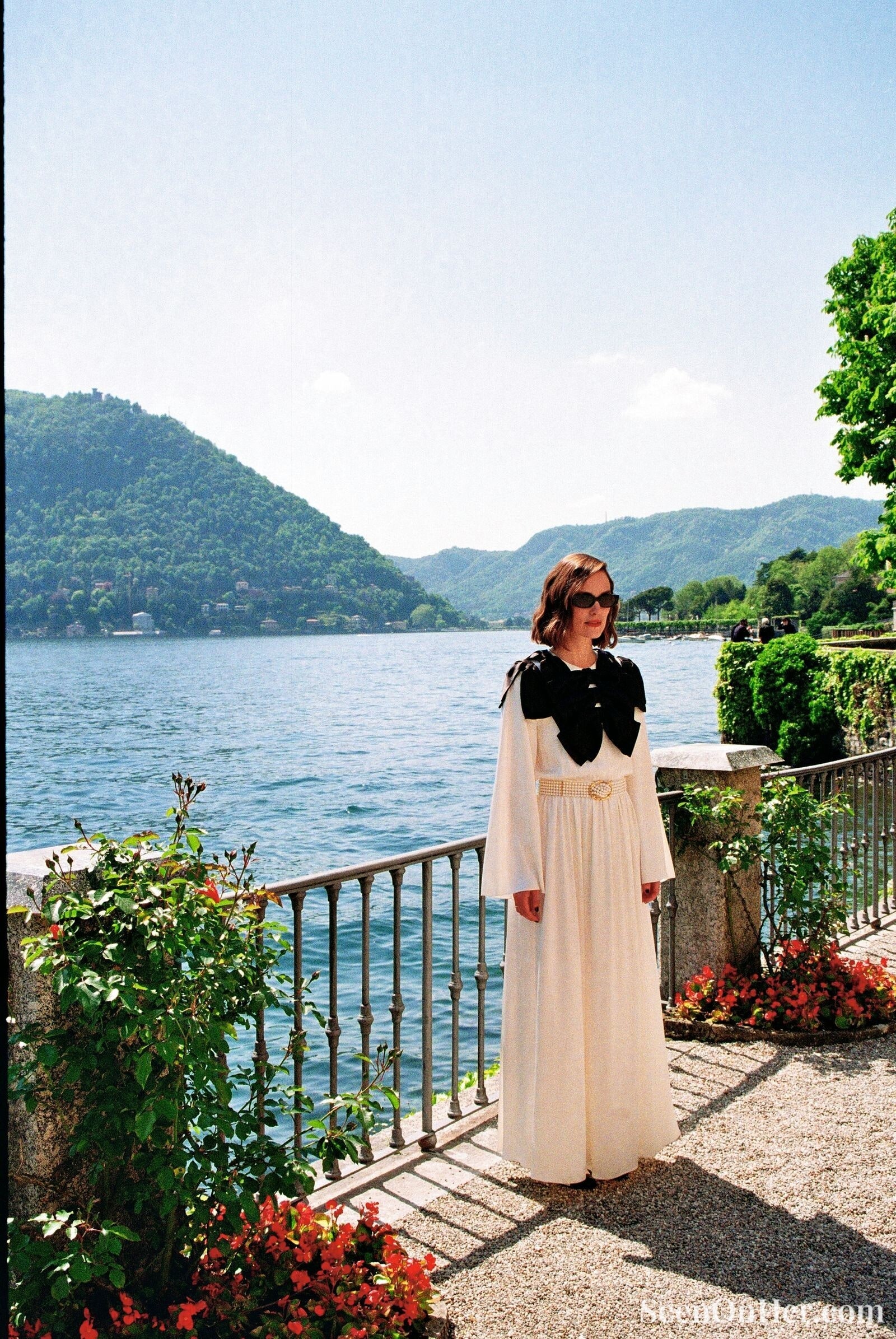 Full-body shot of Keira Knightley in an elegant Chanel gown beside Lake Como, surrounded by colorful flowers and lush greenery, captured in natural sunlight.
