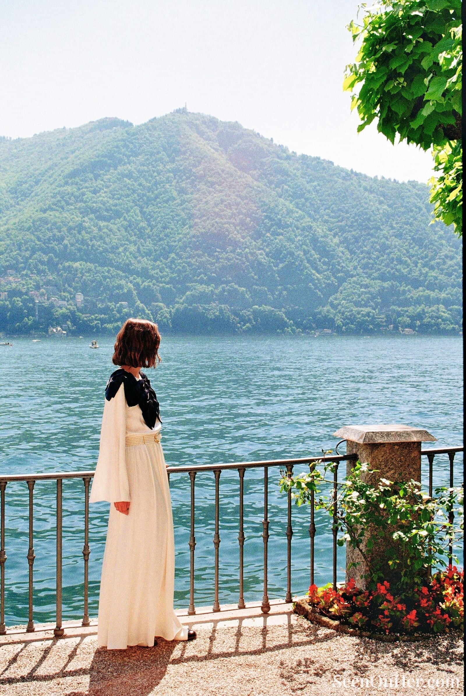 Keira Knightley gazes out at the lake and mountains in an ivory floor-length Chanel dress with pearl belt and black satin detailing, framed by flowers and railing.