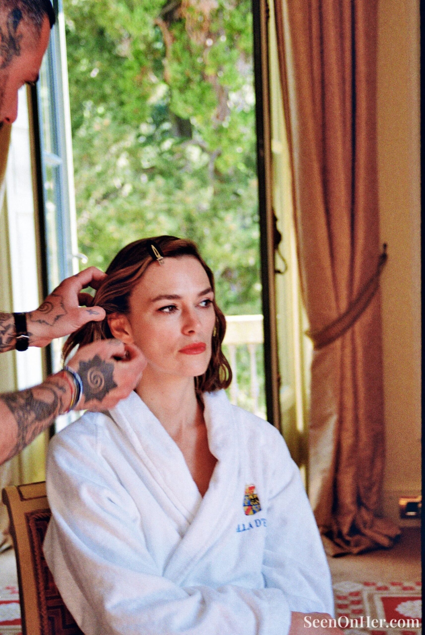 Behind-the-scenes moment of Keira Knightley getting her hair styled in a white hotel robe before the Chanel Cruise 2025 show, seated near an open balcony.