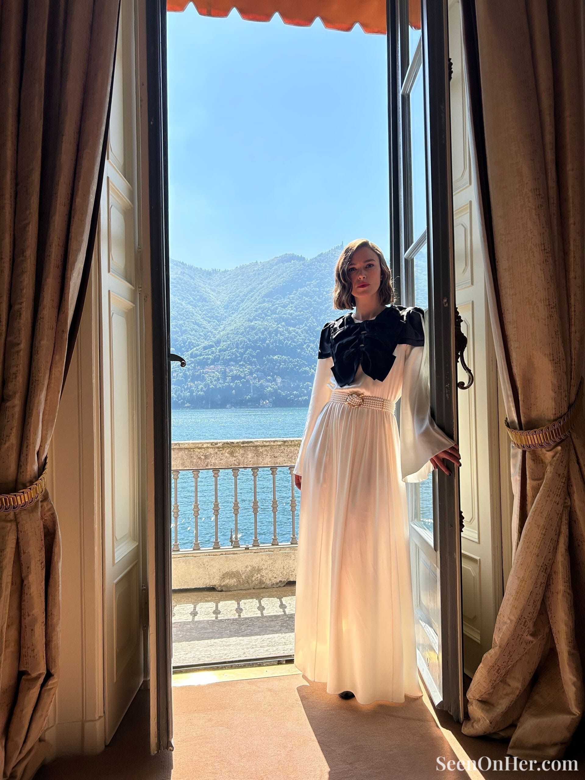 Keira Knightley stands in a sunlit doorway overlooking Lake Como, wearing a flowing white Chanel dress with black bows, framed by elegant curtains and blue skies.