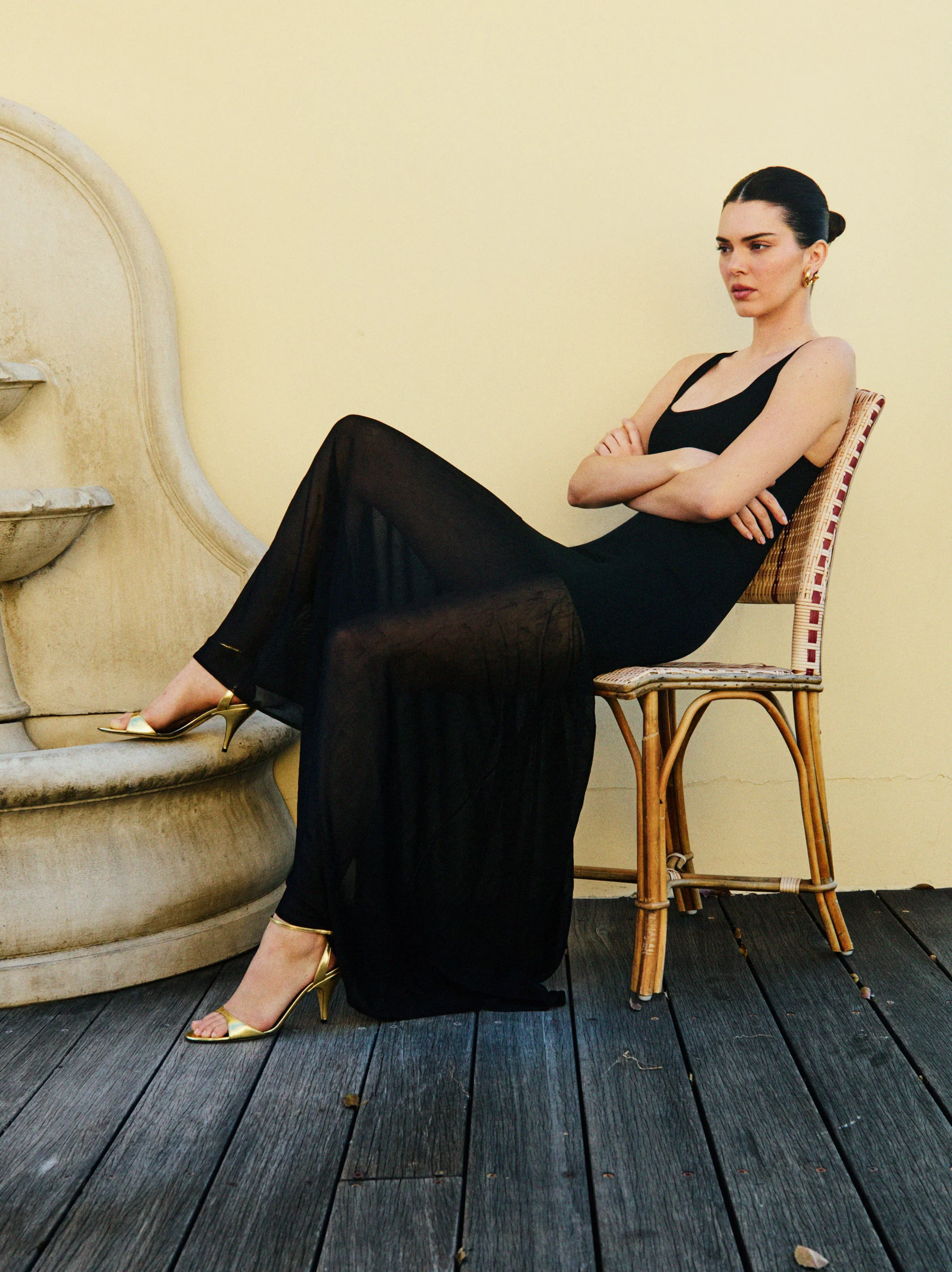 Kendall Jenner sitting on a patio chair in a sheer black maxi dress and gold heels next to a fountain for FWRD Spring 2025