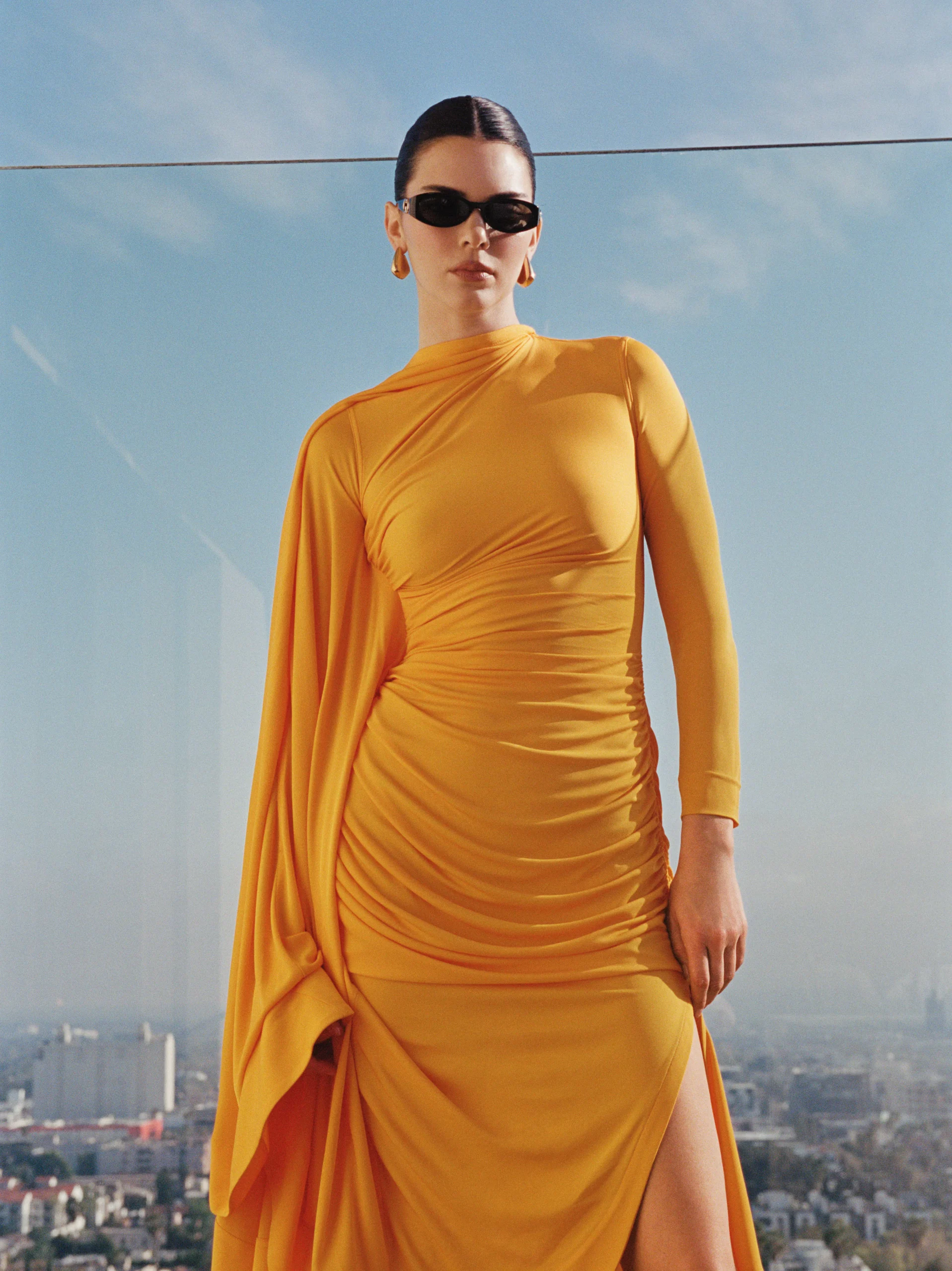Kendall Jenner in a bold yellow ruched gown with high slit and sunglasses, posing against a city skyline backdrop for FWRD Spring 2025
