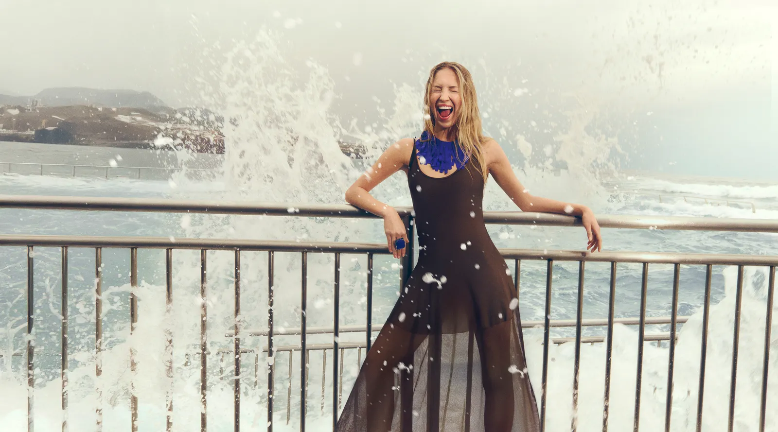Lila Moss laughs while standing in front of crashing ocean waves, dressed in a sheer black dress with a bold blue necklace.