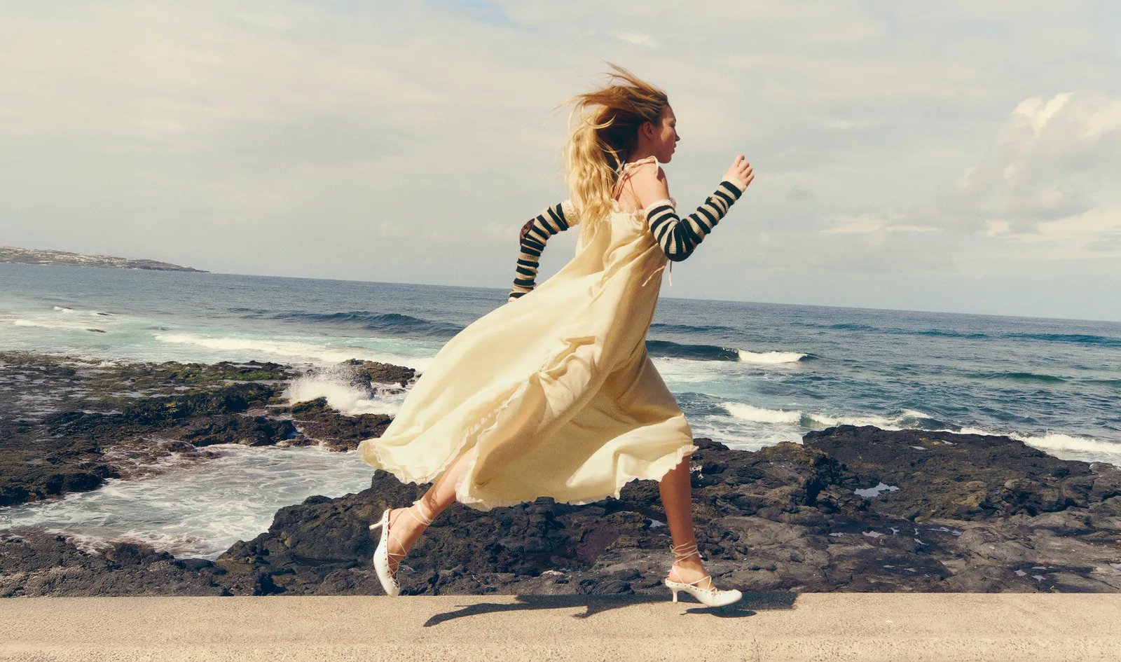 Lila Moss running along a rocky coastal path in a flowing cream dress and striped sleeves for Vogue Spain April 2025