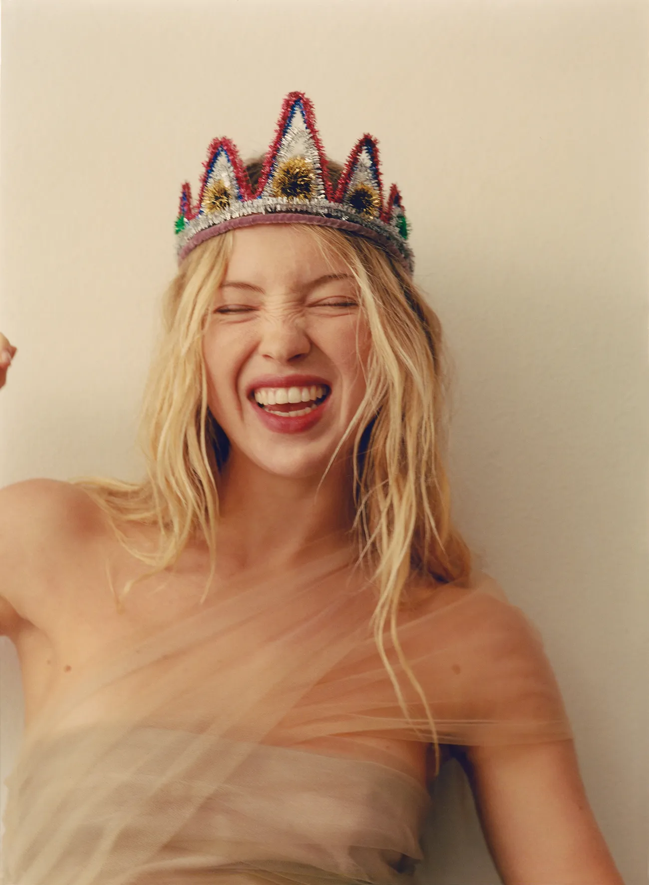 Lila Moss playfully smiles wearing a sheer nude dress and a colorful beaded crown, shot against a neutral background.