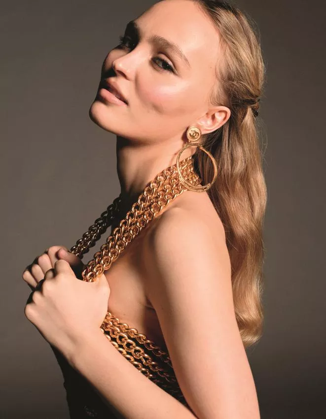 Close-up of Lily-Rose Depp wearing chunky gold chain jewelry and Chanel earrings, with sleek side-parted waves.