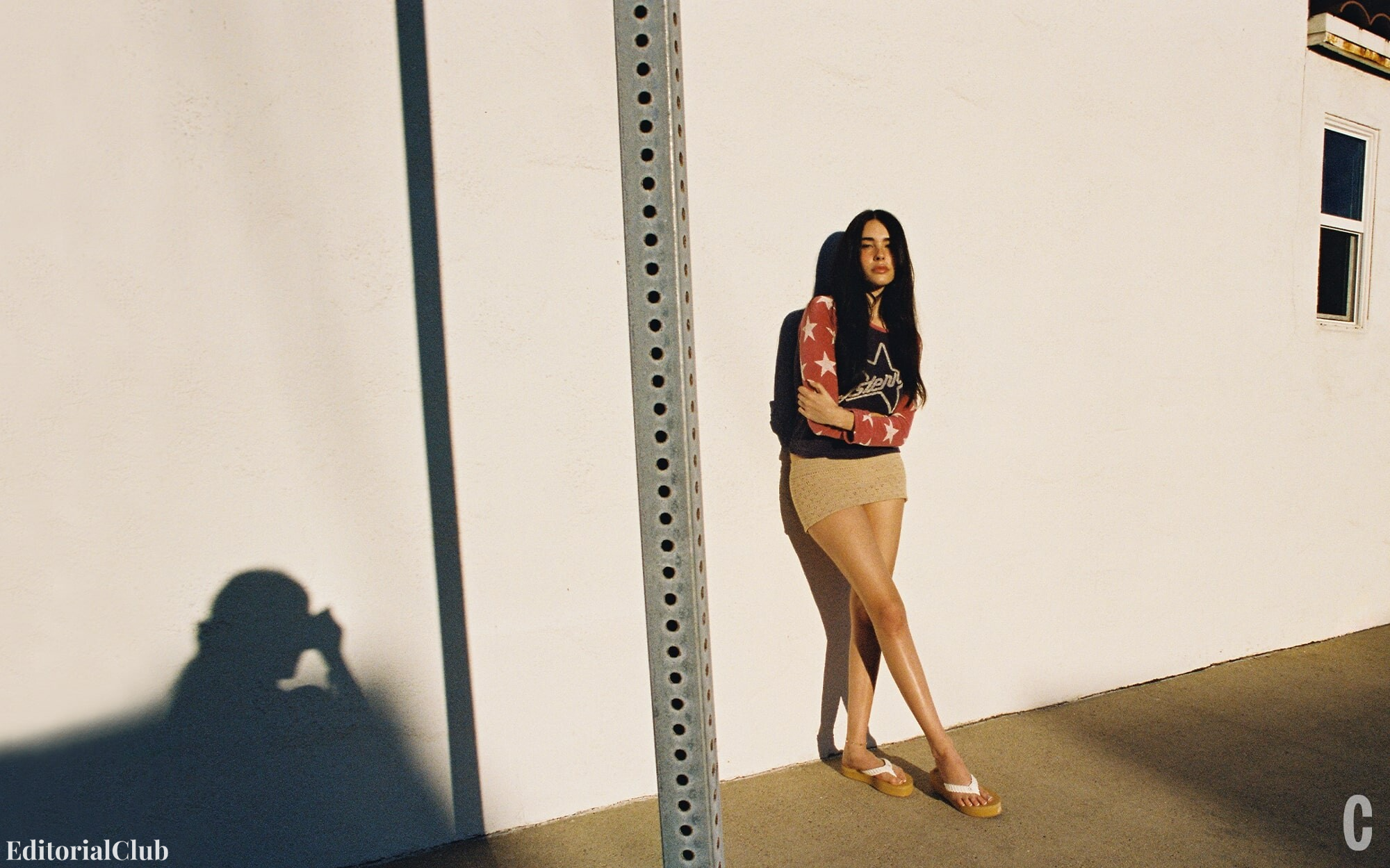 Madison Beer leaning against a white wall in a mini skirt and a red star-print top, with the shadow of a photographer in the foreground.