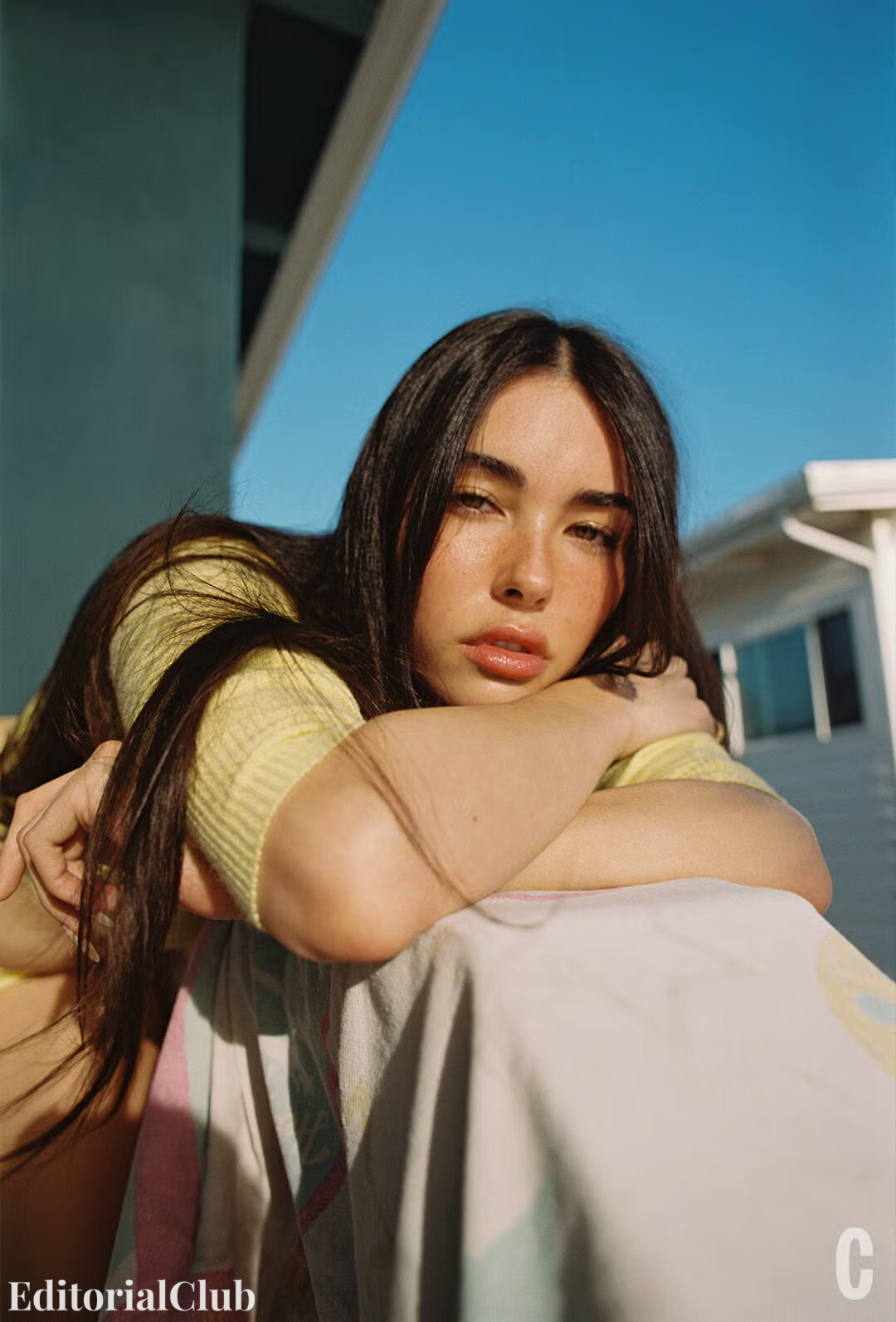 Madison Beer resting her head on her arms while sitting outdoors, wearing a soft yellow sweater and gazing into the camera.