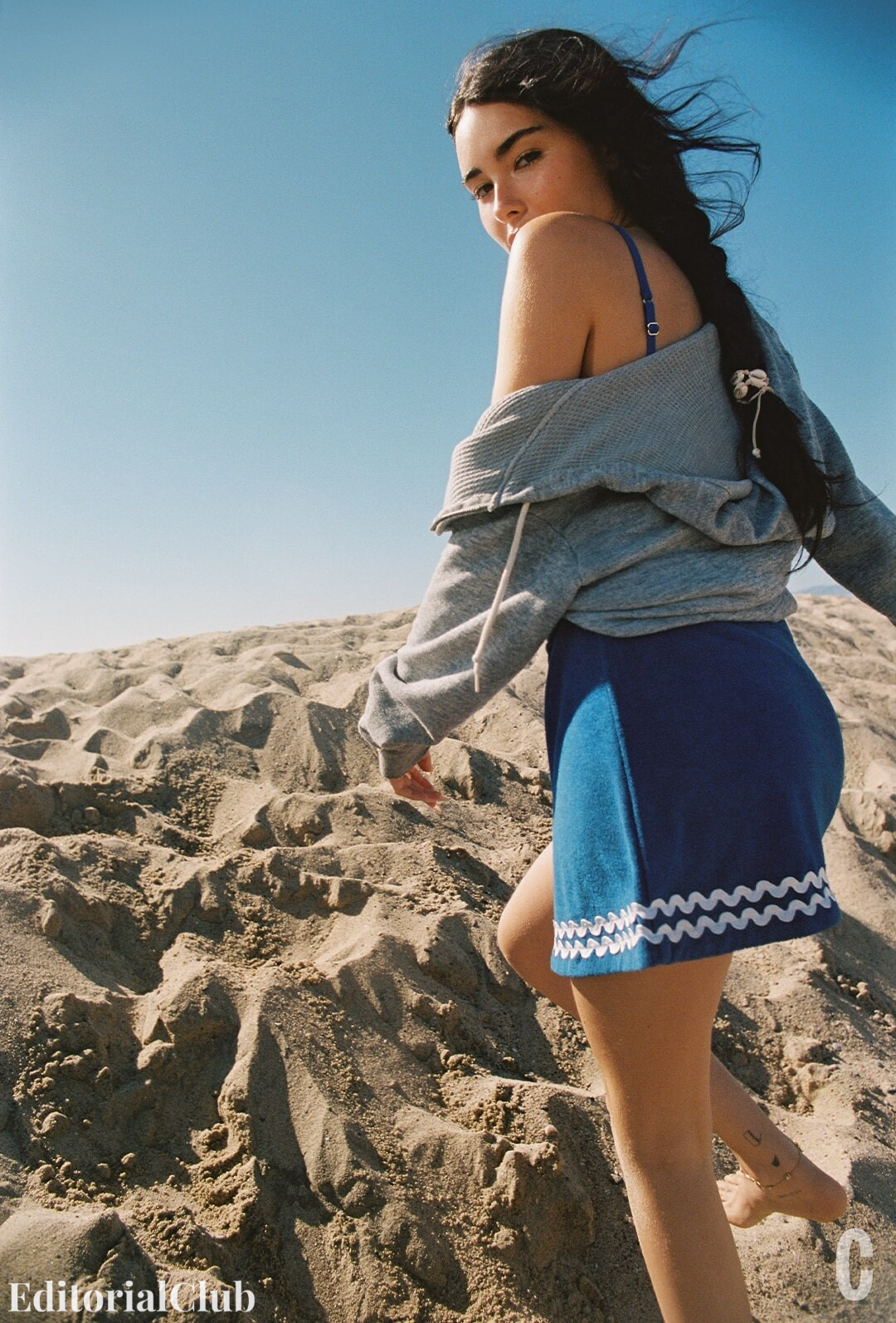 Madison Beer walking up a sand dune in a blue mini skirt and grey hoodie, with wind blowing through her braided hair.