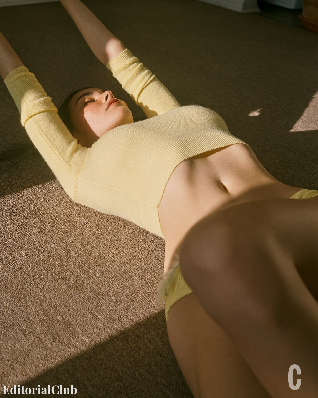 Madison Beer stretching on a carpeted floor in a yellow crop top and matching bottoms, basking in natural sunlight.