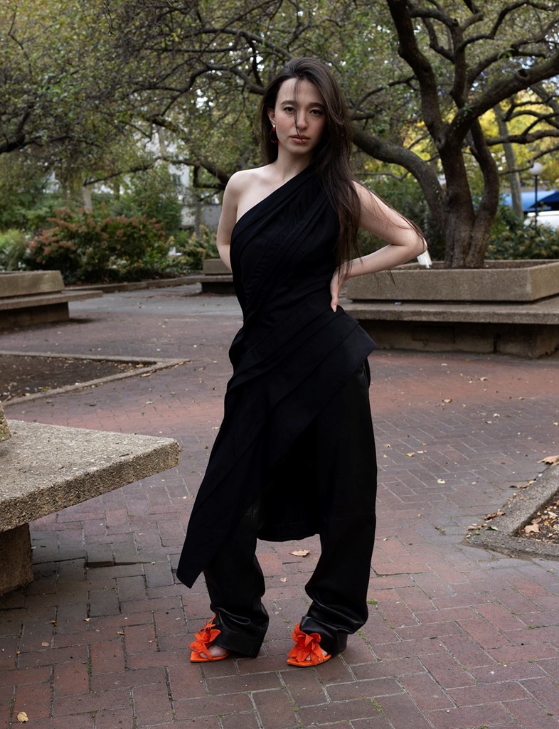 Mikey Madison in an asymmetrical black outfit with orange heels, posing confidently in a Brooklyn park during the Dazed Winter 2025 shoot.