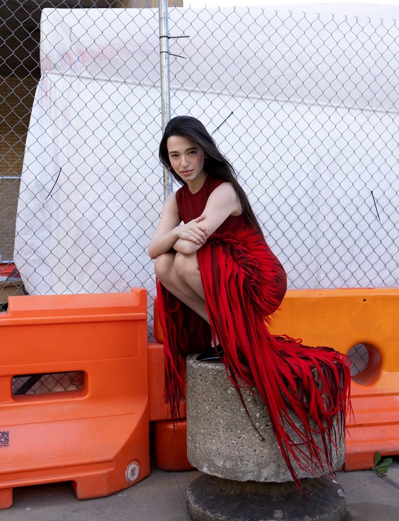 Mikey Madison crouched on a concrete post wearing a dramatic red fringe dress, shot against an urban construction backdrop in Brooklyn.