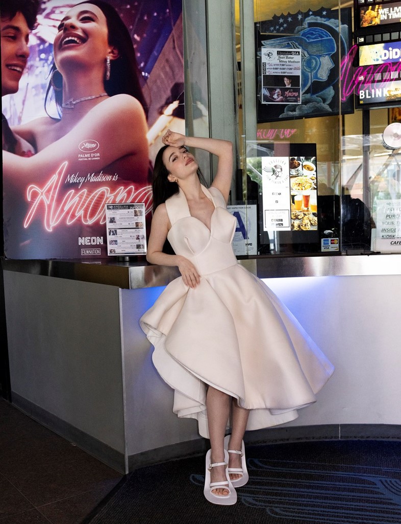 Mikey Madison in a sculptural white cocktail dress and platform sandals, posing at a movie theater lobby in front of an “Anora” film poster.