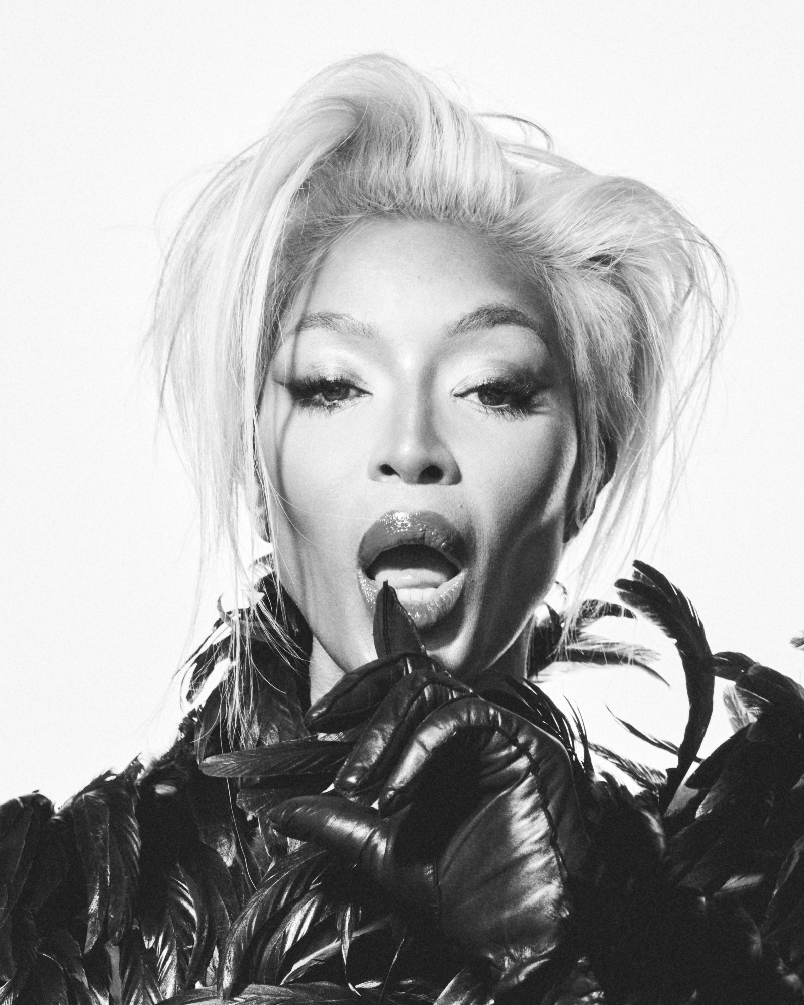 Naomi Campbell in a feathered outfit with bold makeup, striking a playful expression with gloved hands in a high-fashion black and white portrait.
