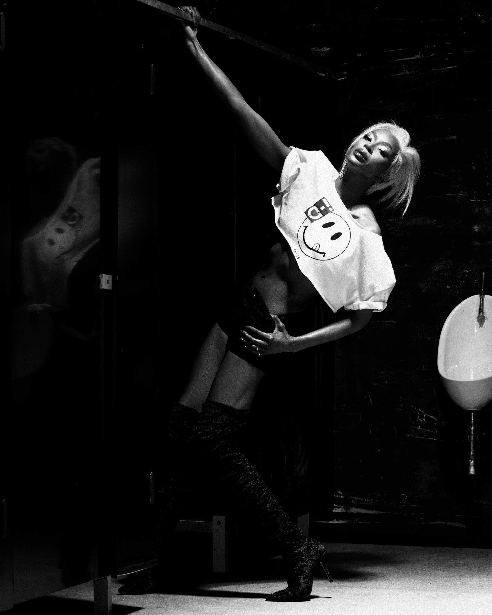 Naomi Campbell posing in a cropped smiley-face tee and velvet boots, leaning in a dramatic pose inside a dark restroom for i-D Magazine.