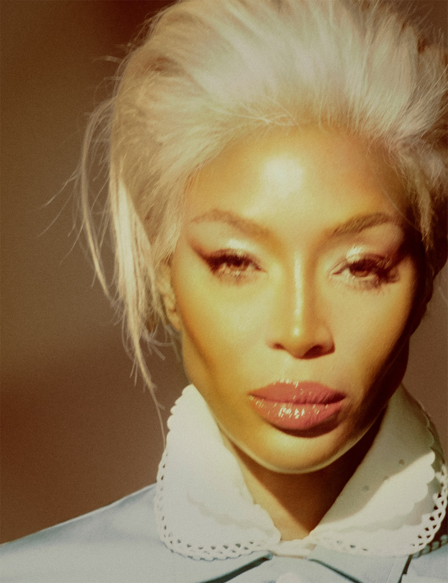 Close-up portrait of Naomi Campbell with platinum blonde hair and glossy lips, wearing a pale blue collared top for i-D Magazine March 2025.