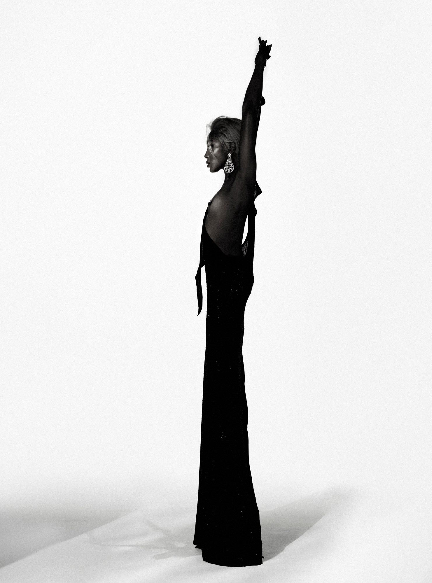 Side profile of Naomi Campbell in a backless gown with arms raised, wearing chandelier earrings in a monochrome i-D Magazine March 2025 image.