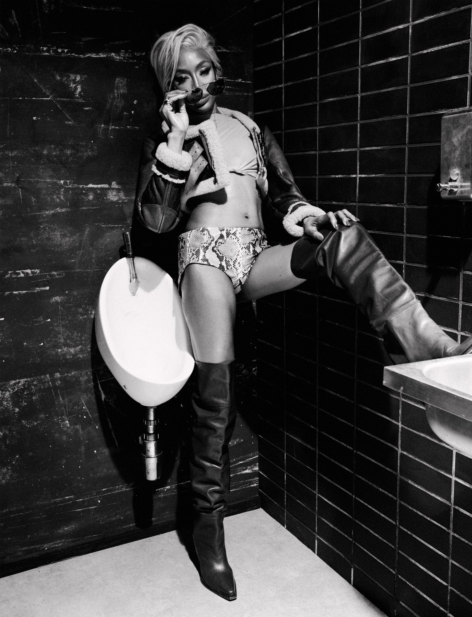 Naomi Campbell leans against a urinal in a grunge restroom wearing a snakeskin bikini bottom, crop top, aviator jacket, and thigh-high boots for i-D Magazine March 2025.