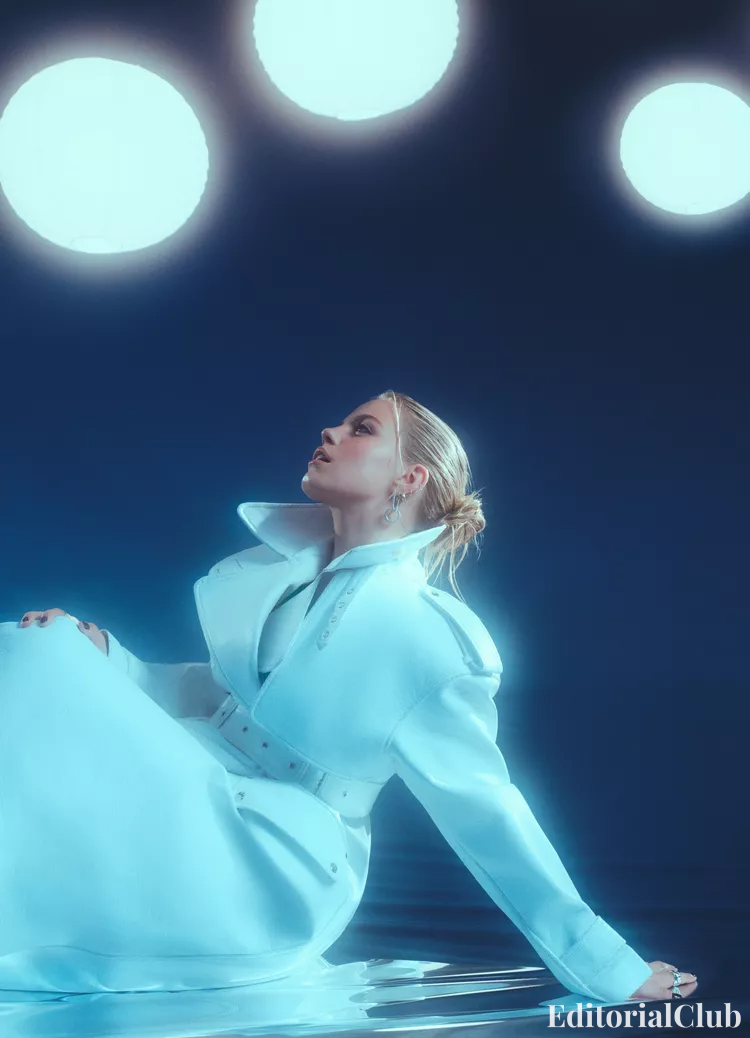 Side profile of Renee Rapp in a structured icy blue coat with dramatic collar, gazing upwards against blue backdrop