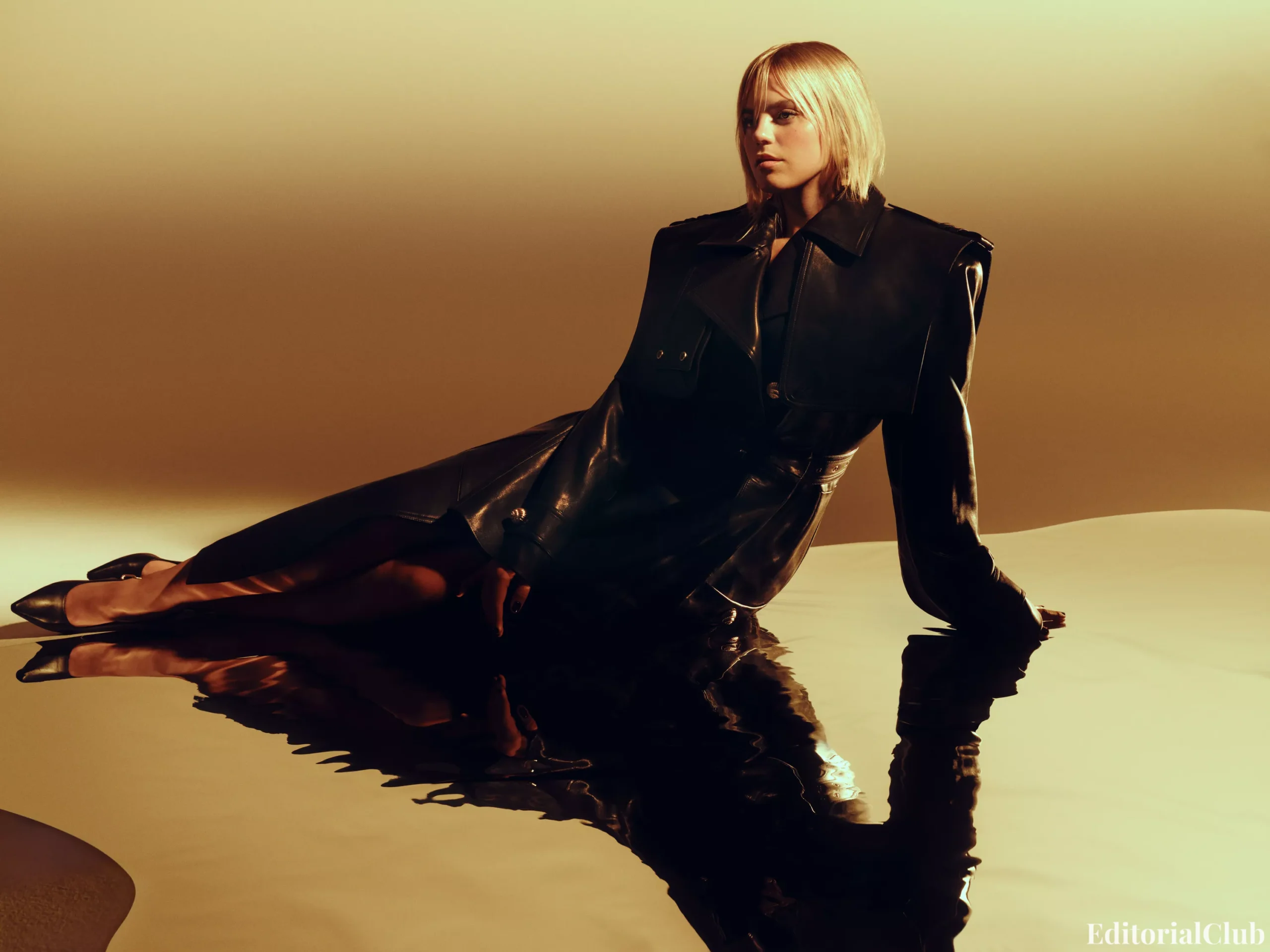 Renee Rapp seated in a black leather trench coat with pointed heels, reflected on glossy floor for InStyle magazine