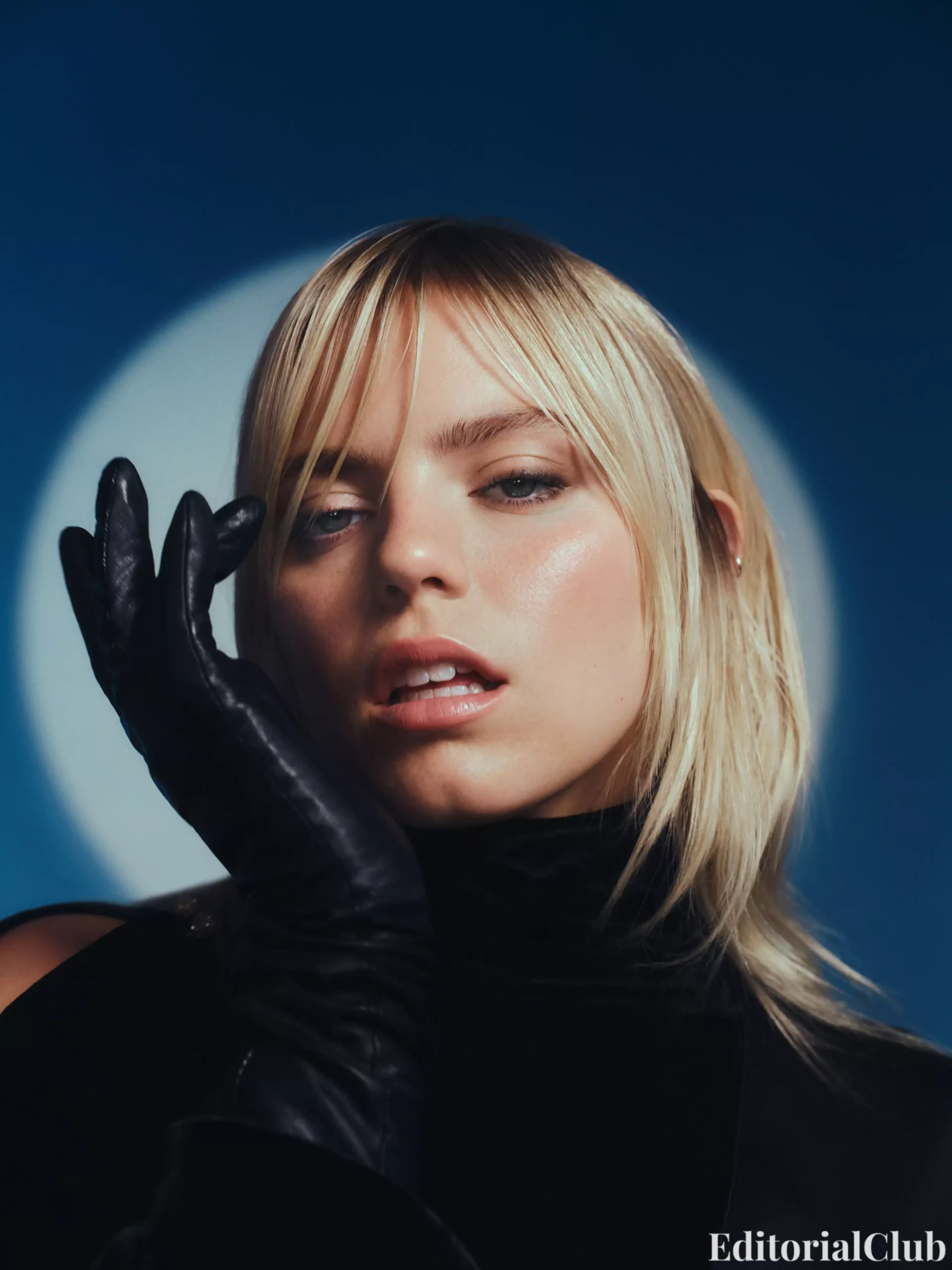Close-up of Renee Rapp wearing black gloves, gently touching her face in moody blue lighting