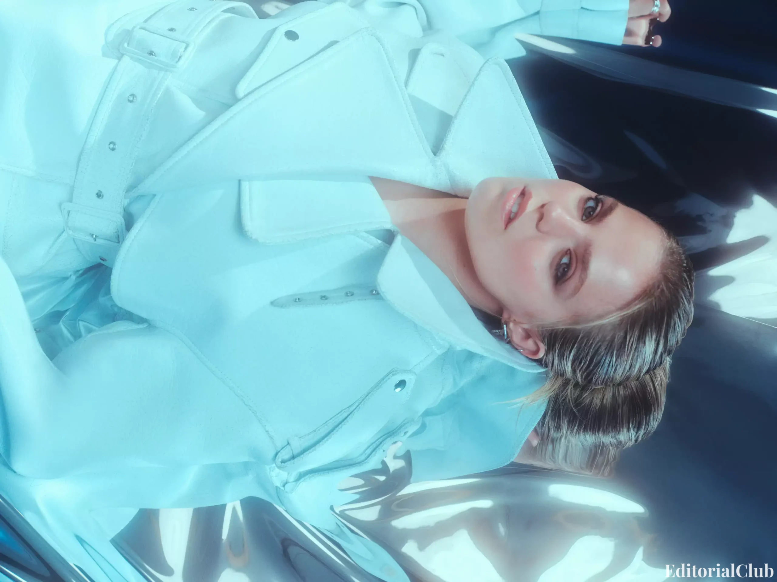 Renee Rapp lying down in a pale blue belted coat, styled with slicked-back hair and glossy background for InStyle