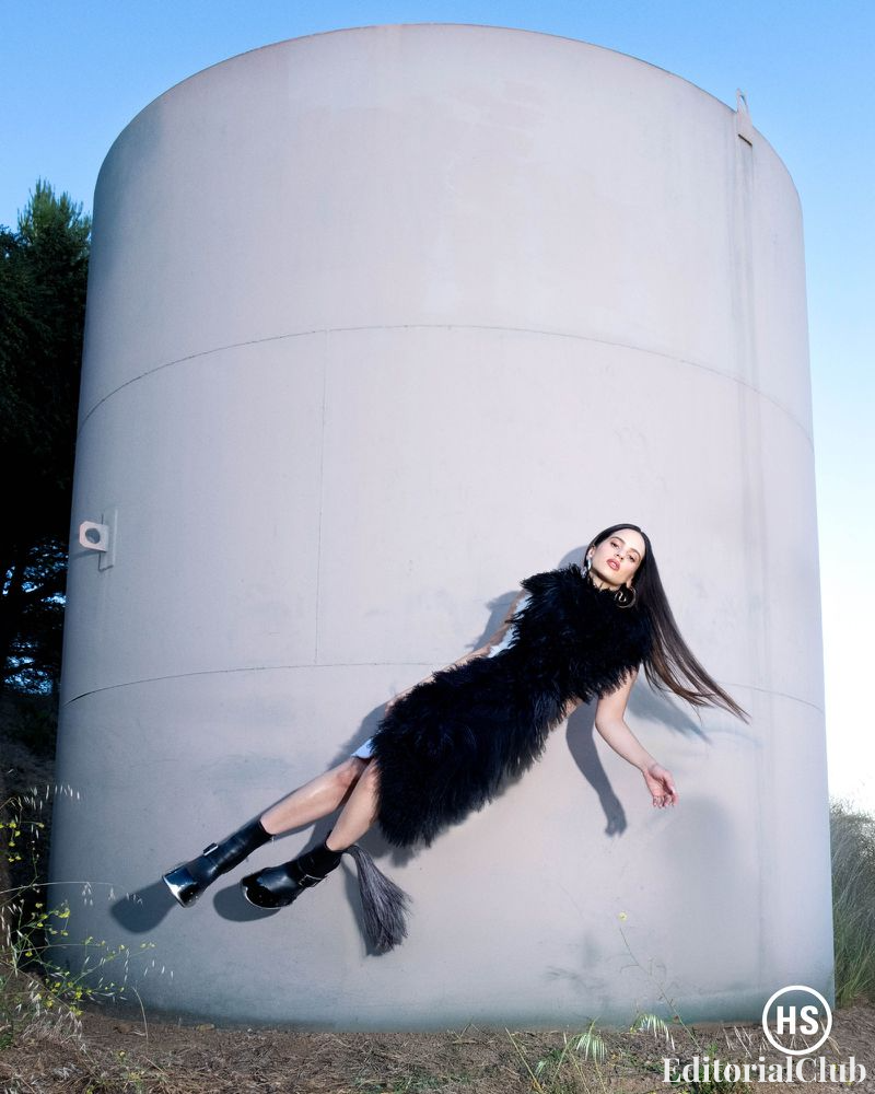 Rosalía wearing a black fur dress, appearing to float horizontally against a curved concrete wall at sunset, for Highsnobiety Fall 2024.