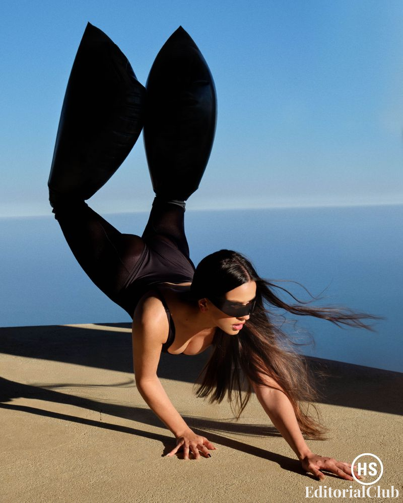 Rosalía in an all-black bodysuit with exaggerated leg accessories and futuristic sunglasses, striking a dynamic pose over a cliffside with ocean view.