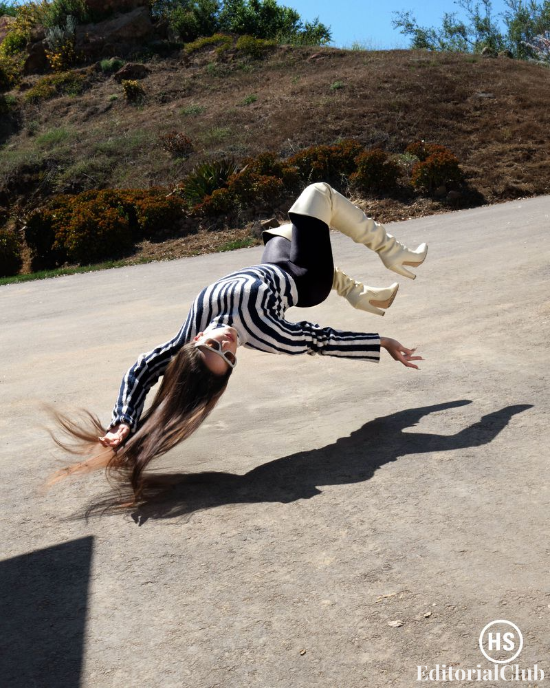 Rosalía mid-flip in a bold striped sweater and white boots, defying gravity on a sunlit slope surrounded by nature.