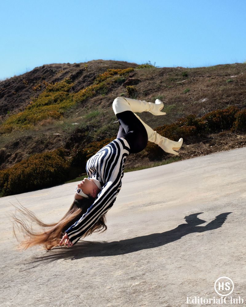 Rosalía twisting midair in striped top and white boots, hair suspended dramatically during outdoor shoot.