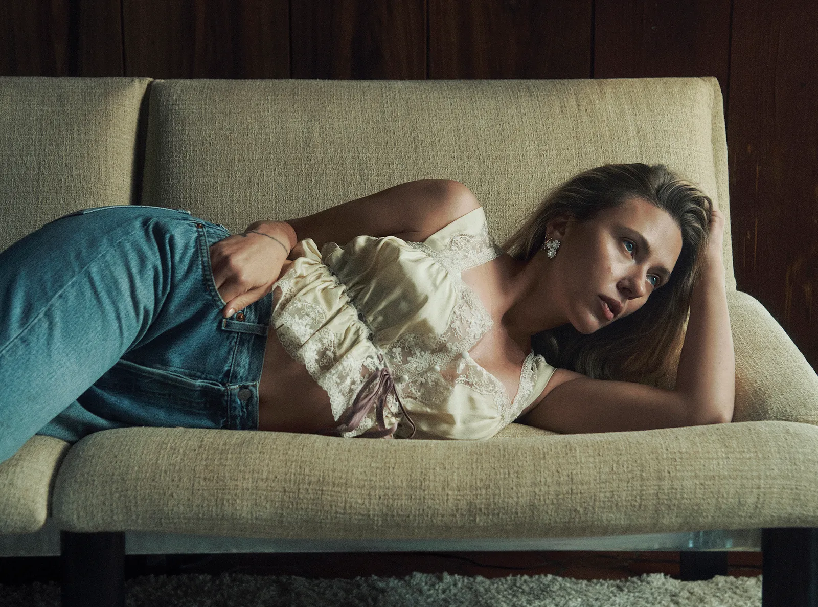 Scarlett Johansson lying on a vintage beige sofa, wearing jeans and a lacy ivory camisole with vintage earrings.