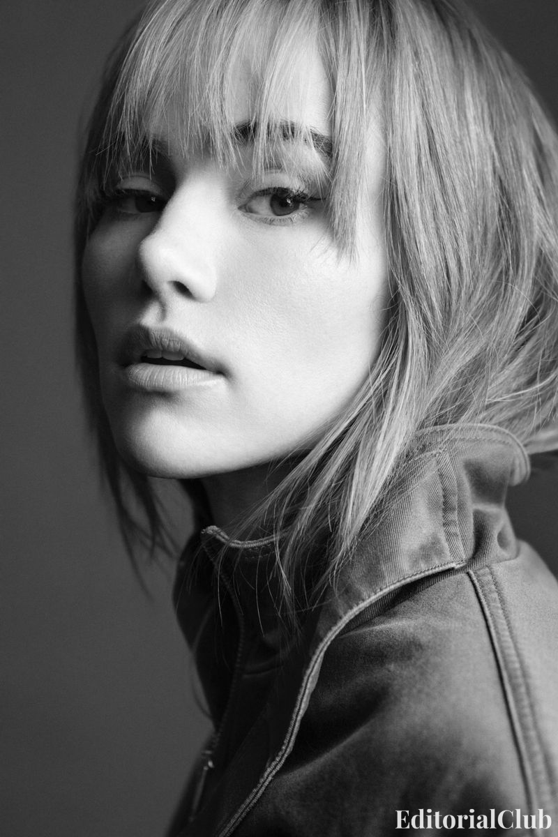 Black and white close-up portrait of Suki Waterhouse with tousled blonde bangs and a zipped jacket, gazing intensely sideways.