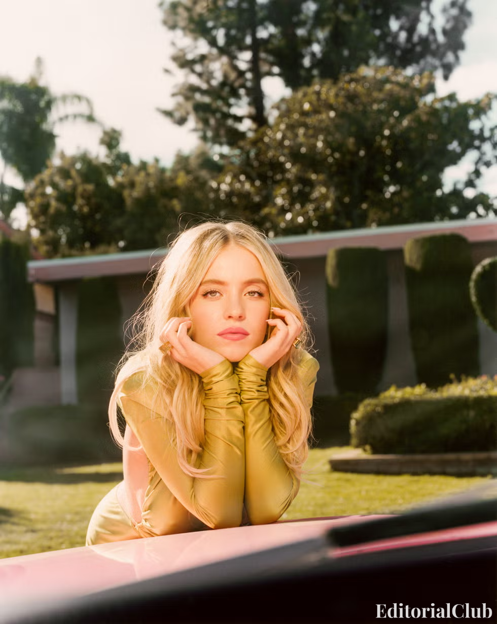 Sydney Sweeney resting her face on her hands while leaning on a pink car in a dreamy outdoor Cosmopolitan 2022 fashion editorial.